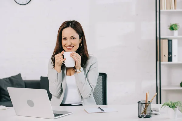 Feliz jovem secretário segurando xícara de café e olhando para a câmera enquanto sentado no local de trabalho no escritório — Fotografia de Stock