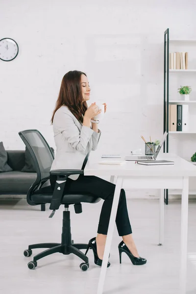 Elegante, junge Sekretärin sitzt mit geschlossenen Augen am Arbeitsplatz und hält eine Kaffeetasse in der Hand — Stockfoto