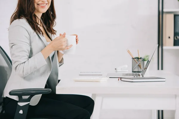 Обрезанный вид улыбающейся секретарши, держащей чашку кофе на рабочем месте в офисе — стоковое фото