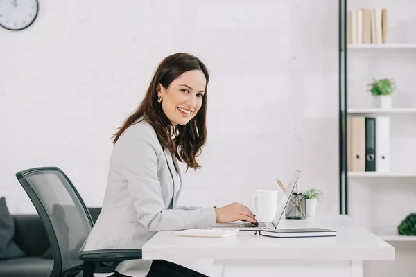 Joven, secretaria sonriente mirando a la cámara mientras está sentado en el lugar de trabajo en la oficina - foto de stock