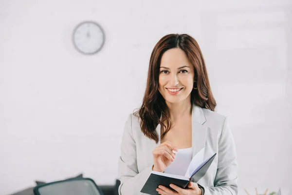 Hermosa, secretaria sonriente mirando a la cámara mientras escribe en el cuaderno - foto de stock