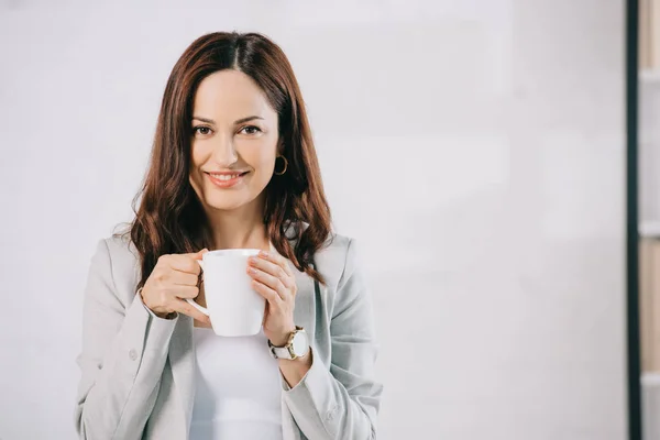 Atraente, jovem secretária olhando para a câmera enquanto segurando xícara de café — Fotografia de Stock