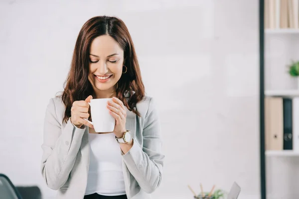 Attraktiv, junge Sekretärin lächelt, während sie eine Kaffeetasse im Büro hält — Stockfoto