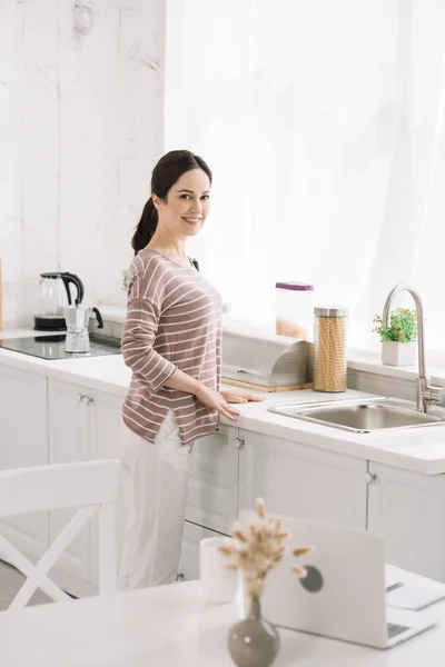 Селективный фокус кухонного стола с ноутбуком рядом с улыбающейся женщиной, стоящей возле раковины — стоковое фото