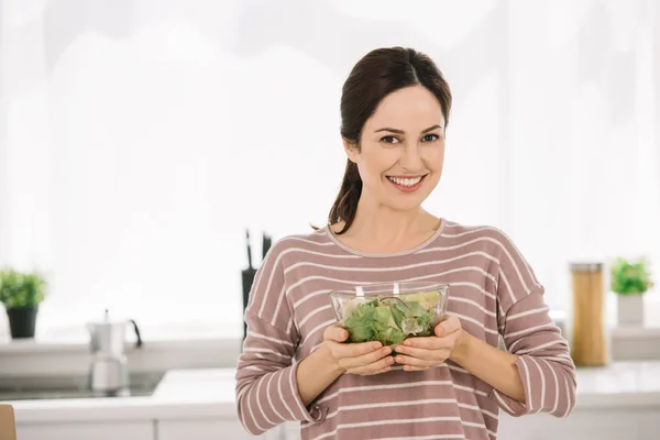 Glückliche junge Frau lächelt in die Kamera, während sie eine Schüssel mit frischem Gemüsesalat in der Hand hält — Stockfoto