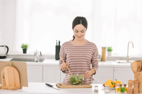 Jovem, mulher atraente misturando salada de legumes frescos enquanto está na mesa da cozinha — Fotografia de Stock