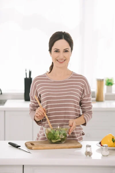 Счастливая молодая женщина смотрит в камеру, смешивая свежий овощной салат — стоковое фото