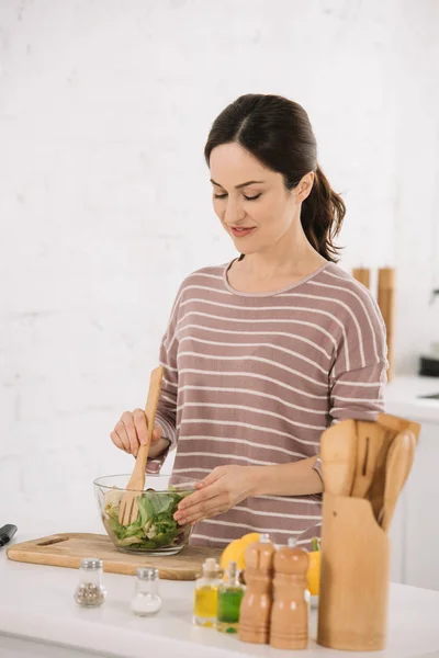 Beautiful young woman mixing fresh vegetable salad while standing at kitchen table — Stock Photo