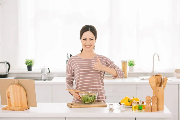 Feliz joven mostrando el pulgar hacia arriba mientras está de pie cerca de un tazón con ensalada de verduras frescas - foto de stock