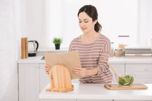 Atractiva y sonriente mujer leyendo el libro de recibos mientras está de pie en la mesa de la cocina cerca del tazón con ensalada de verduras - foto de stock