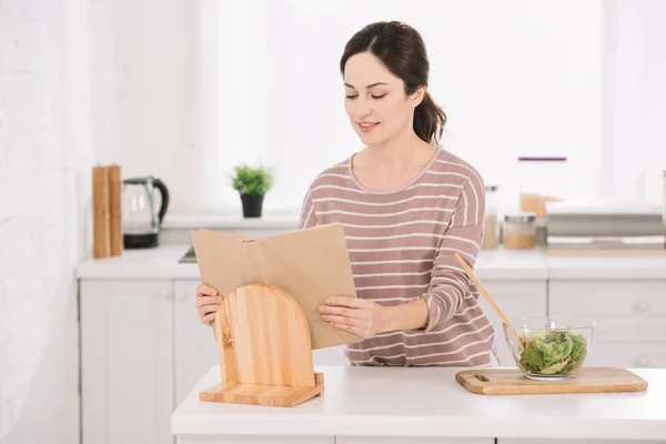Attractive, smiling woman reading receipt book while standing at kitchen table near bowl with vegetable salad — Stock Photo