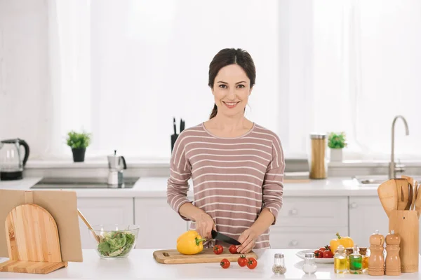 Joven, mujer feliz cortando verduras frescas en la tabla de cortar y mirando a la cámara - foto de stock