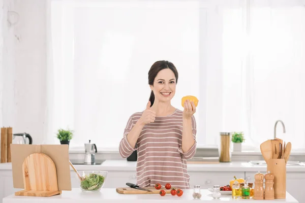 Joven, mujer alegre mostrando el pulgar hacia arriba mientras sostiene el pimiento cortado y mirando a la cámara - foto de stock