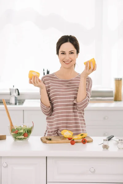 Весела молода жінка дивиться на камеру, стоячи біля кухонного столу зі свіжими овочами і тримаючи нарізаний болгарський перець — стокове фото