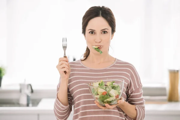 Attraente giovane donna guardando la fotocamera mentre tiene la forchetta e mangiare insalata di verdure fresche — Foto stock