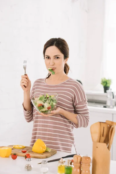 Bella giovane donna guardando la fotocamera mentre tiene la forchetta e mangiare insalata di verdure fresche — Foto stock