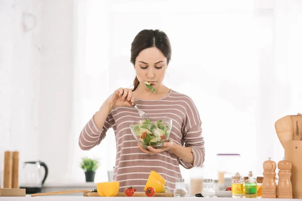 Atraente, jovem mulher comer salada de legumes enquanto está em pé na mesa da cozinha com legumes frescos — Fotografia de Stock