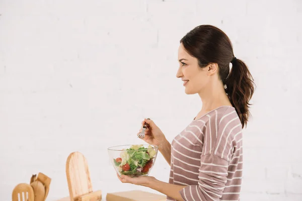 Side view of attractive, smiling woman holding bowl with fresh vegetable salad — Stock Photo