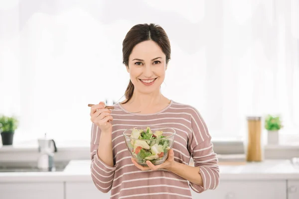 Attraente, donna sorridente guardando la fotocamera mentre tiene ciotola con insalata di verdure — Foto stock