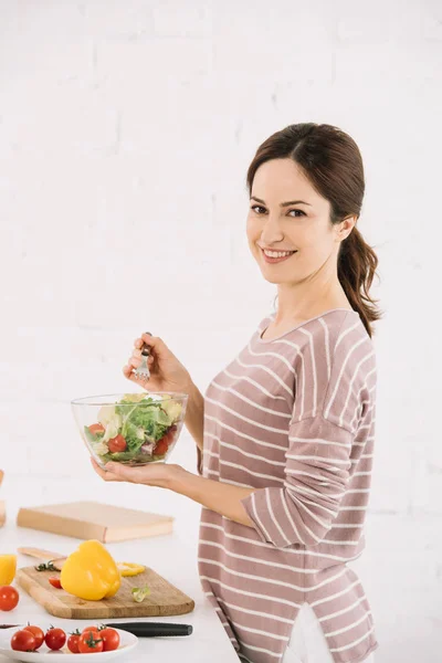 Pretty, cheerful woman looking at camera while holding bowl with fresh vegetable salad — Stock Photo