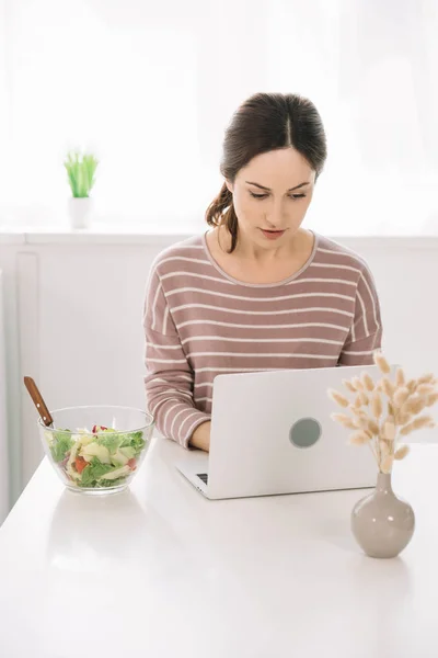 Attraente, donna attenta utilizzando il computer portatile mentre seduto al tavolo della cucina vicino ciotola con insalata di verdure — Foto stock