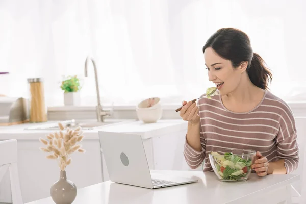 Giovane, donna felice mangiare insalata di verdure mentre seduto al tavolo della cucina vicino al computer portatile — Foto stock