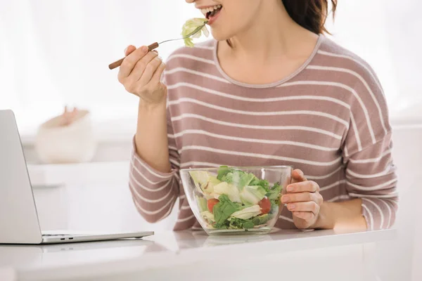 Vista recortada de la joven sentada en la mesa cerca de la computadora portátil y comer ensalada de verduras - foto de stock