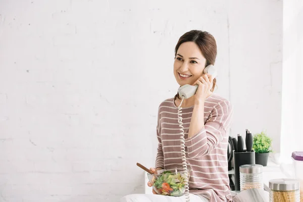 Smiling woman talking on retro phone while sitting on kitchen table and holding bowl with vegetable salad — Stock Photo