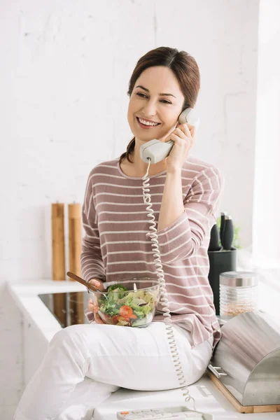 Smiling woman talking on retro phone while sitting on kitchen table and holding bowl with vegetable salad — Stock Photo