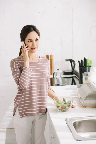 Young, cheerful woman talking on smartphone while standing at kitchen table near bowl with vegetable salad — Stock Photo
