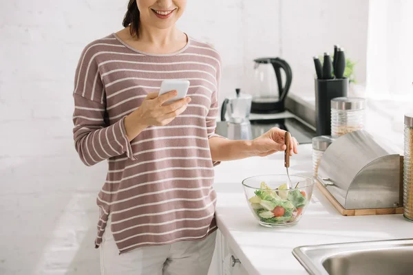 Vue recadrée d'une femme souriante utilisant un smartphone debout à la table de cuisine près d'un bol avec salade de légumes — Photo de stock