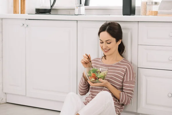 Young, happy woman sitting on floor in kitchen and eating vegetable salad — Stock Photo