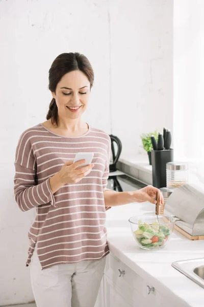Jovem, mulher sorridente usando smartphone enquanto está na mesa da cozinha perto da tigela com salada de legumes — Fotografia de Stock