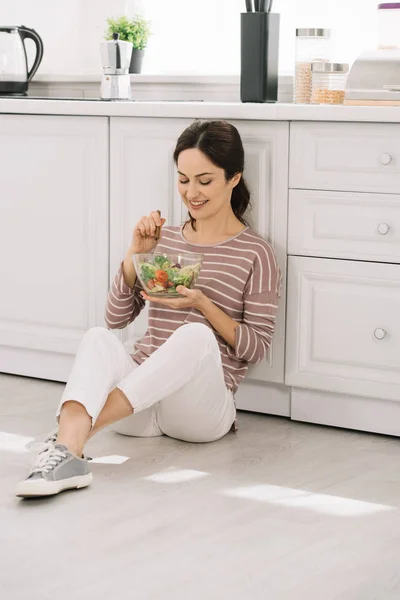 Молодая, веселая женщина сидит на полу на кухне и ест овощной салат — стоковое фото