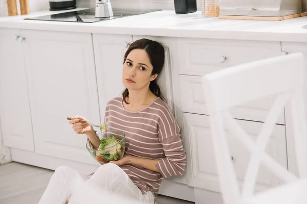 Unzufriedene junge Frau sitzt in Küche auf dem Boden und isst Gemüsesalat — Stockfoto