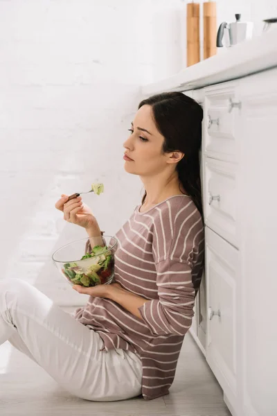 Mujer joven molesta sentada en el suelo en la cocina y comer ensalada de verduras - foto de stock