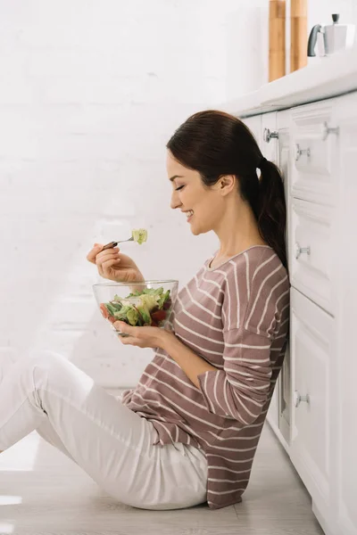 Happy young woman sitting on floor in kitchen and eating vegetable salad — Stock Photo