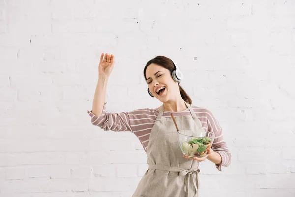 Веселая молодая женщина в наушниках танцует и поет, держа миску с салатом — стоковое фото