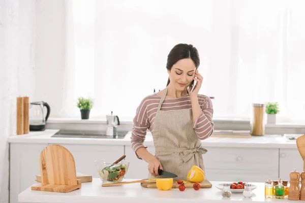 Mujer sonriente en delantal corte de pimiento mientras está de pie cerca de la mesa de la cocina con verduras y hablando en el teléfono inteligente - foto de stock
