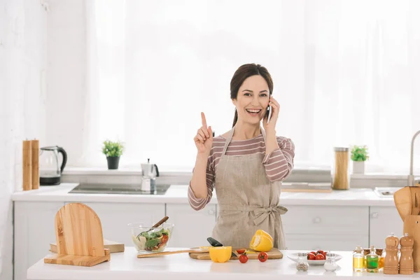 Mujer alegre en delantal mostrando signo de idea mientras está de pie cerca de la mesa de la cocina con verduras y hablando en el teléfono inteligente - foto de stock