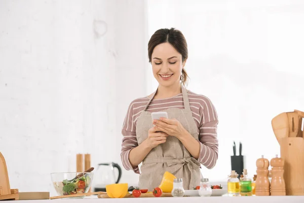 Jeune femme heureuse utilisant smartphone tout en se tenant à la table de cuisine près de légumes frais — Photo de stock