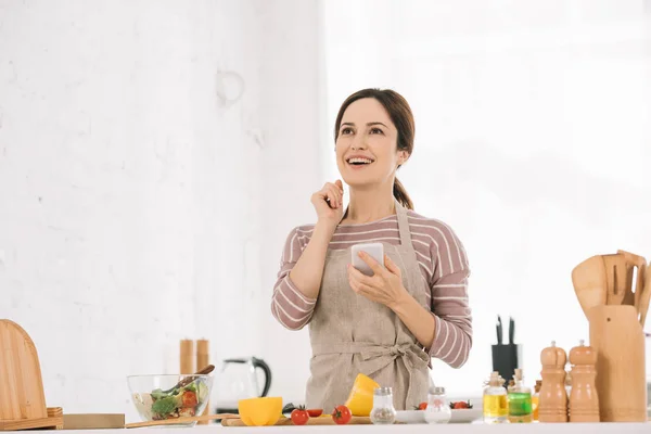 Felice giovane donna in grembiule tenendo smartphone mentre in piedi al tavolo della cucina vicino a verdure fresche — Foto stock