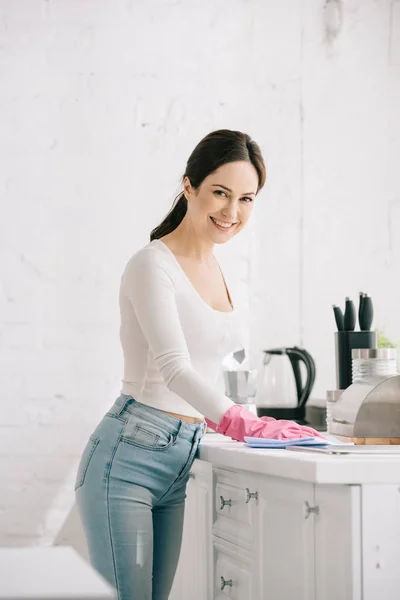 Sorrindo dona de casa olhando para a câmera enquanto limpa a mesa da cozinha com pano — Fotografia de Stock