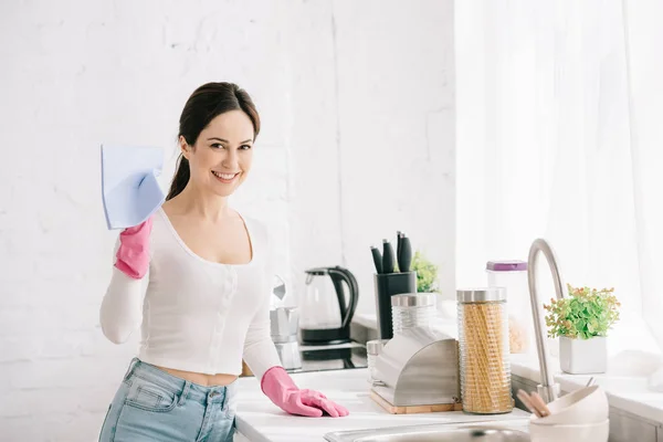 Happy housewife looking at camera while standing in kitchen and holding rag — Stock Photo