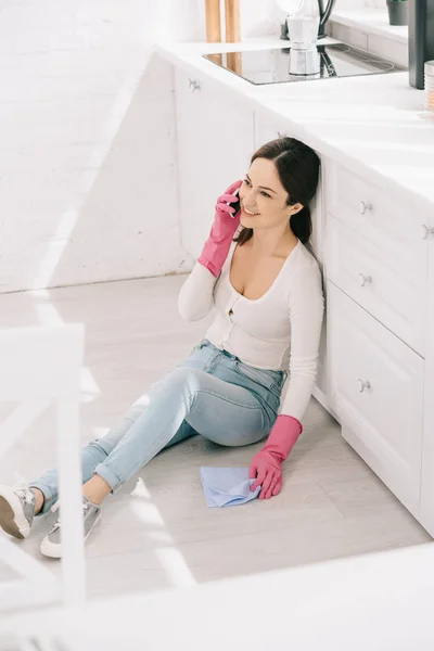 Selective focus of cheerful housewife sitting on floor in kitchen and talking on smartphone — Stock Photo