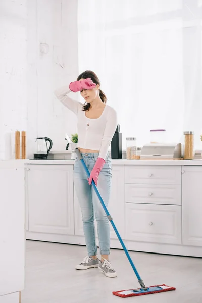 Exhausted housewife standing with mop in kitchen and holding hand near head — Stock Photo