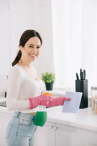 Allegra casalinga guardando la fotocamera mentre tiene bottiglia spray e straccio in cucina — Foto stock