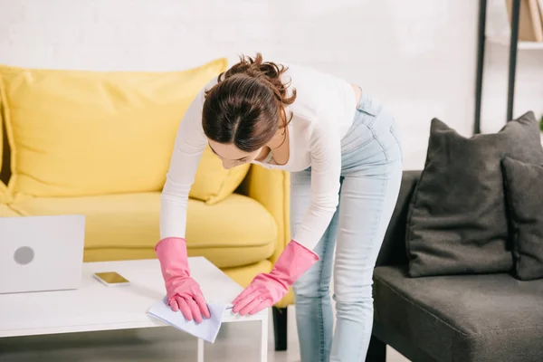 Jovem dona de casa em jeans azul limpando a mesa com pano perto do laptop — Fotografia de Stock