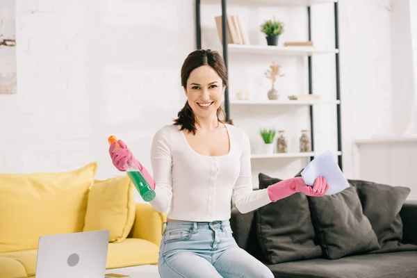 Happy housewife smiling at camera while sitting on table and holding rag and spray bottle — Stock Photo