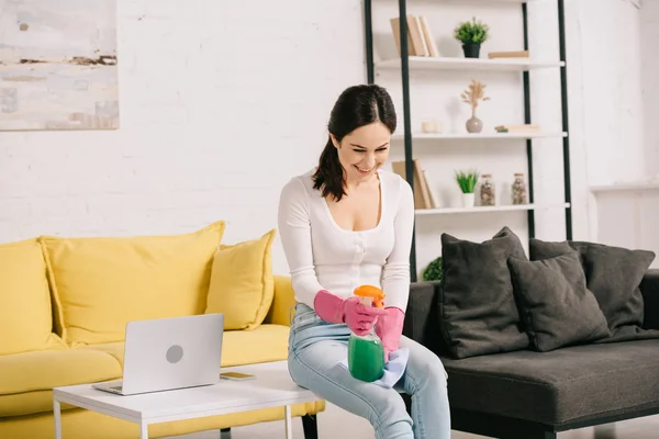 Cheerful housewife smiling while sitting on table and holding rag and spray bottle — Stock Photo